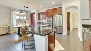 A modern kitchen with a central island, stainless steel appliances, and a dining area with bar stools. - Luxe Family Home w/ Pool, 13 Mi to Dtwn Phoenix! (Laveen)
