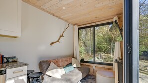 Ein gemütliches Zimmer mit Holzboden, einer Couch mit Kissen, einer kleinen Küchenzeile mit Spüle und Barhocker und einem großen Fenster mit Blick ins Freie.