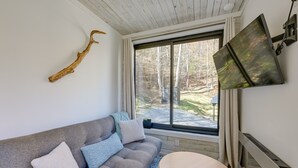 A modern living room with a grey sofa, a round wooden coffee table, a mounted flat-screen TV, and a large window with a forest view. - Bryson City Tiny Cabin w/ Deck Near River Access! (Bryson City)