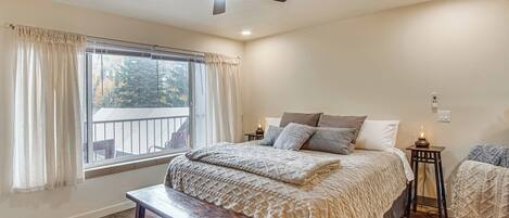 Un dormitorio con una cama grande, un ventilador de techo, una ventana con cortinas, un banco y una mesita de noche con lámpara.