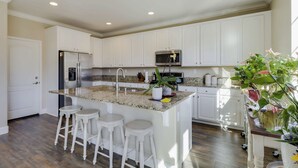 Una cocina con armarios blancos, encimera de granito, refrigerador de acero inoxidable, microondas y un fregadero. Hay tres taburetes blancos y una planta en maceta sobre la encimera.