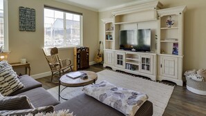 Una sala de estar con un sofá, una mesa de centro, un televisor sobre un mueble blanco, una guitarra y una estantería.