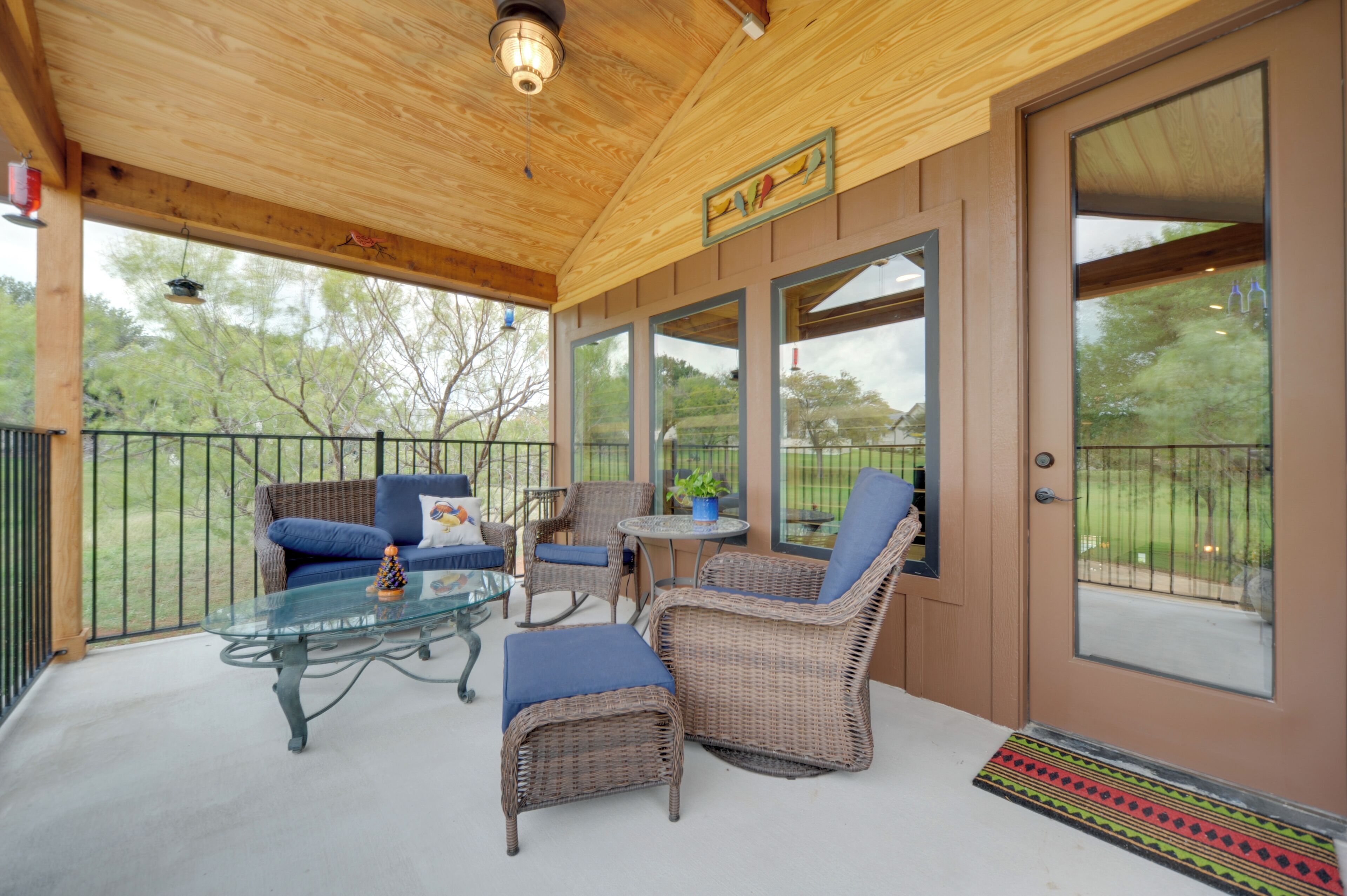 A covered patio with wicker furniture, a glass table, and a view of the outdoors.