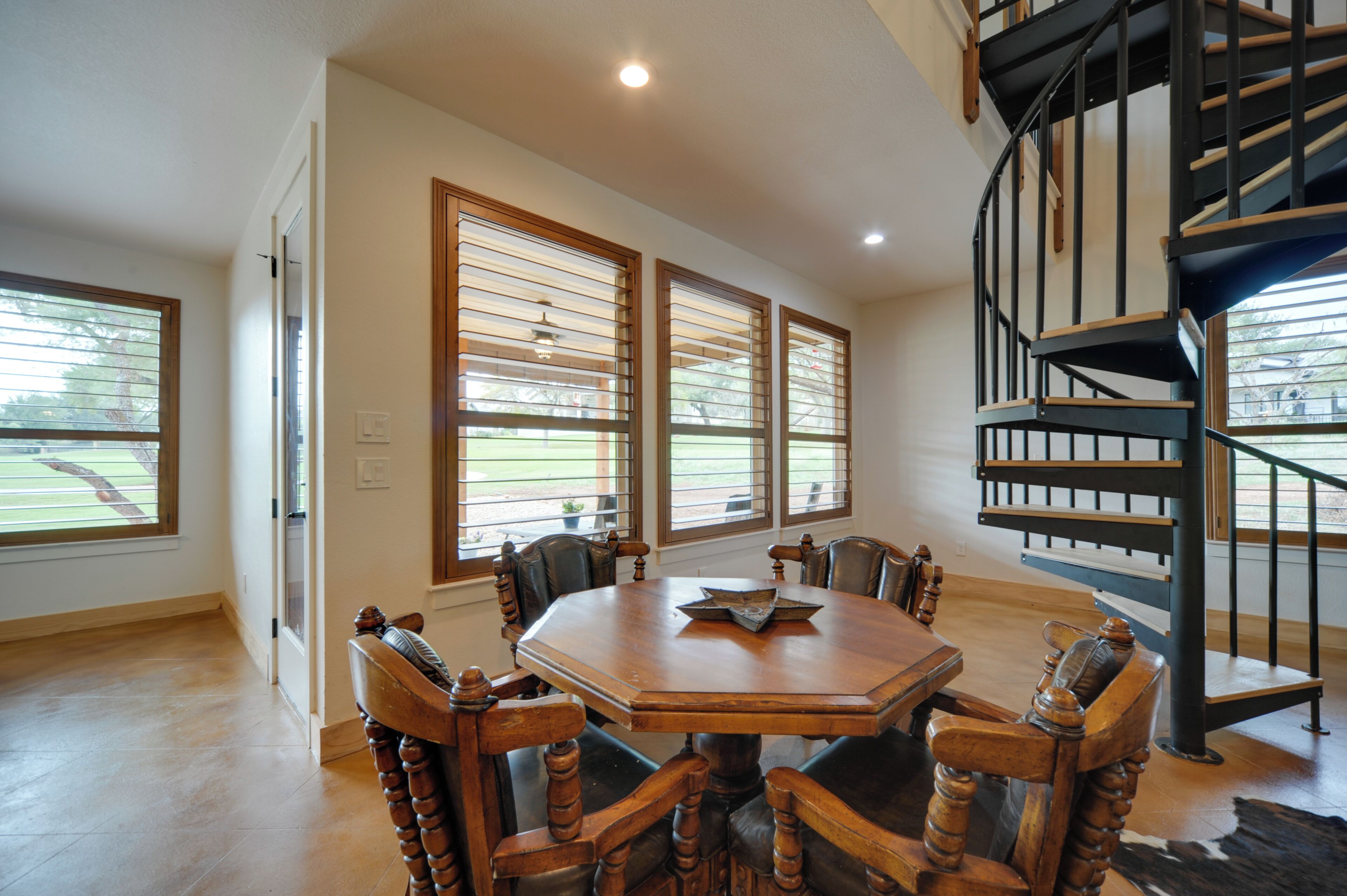 A dining area with a wooden table and chairs, large windows with blinds, and a spiral staircase.