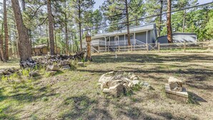 A rustic cabin with a porch, surrounded by tall pine trees and a fenced yard with rocks and logs. - Rustic Alto Retreat - Near Hiking, Skiing & More! (Alto)