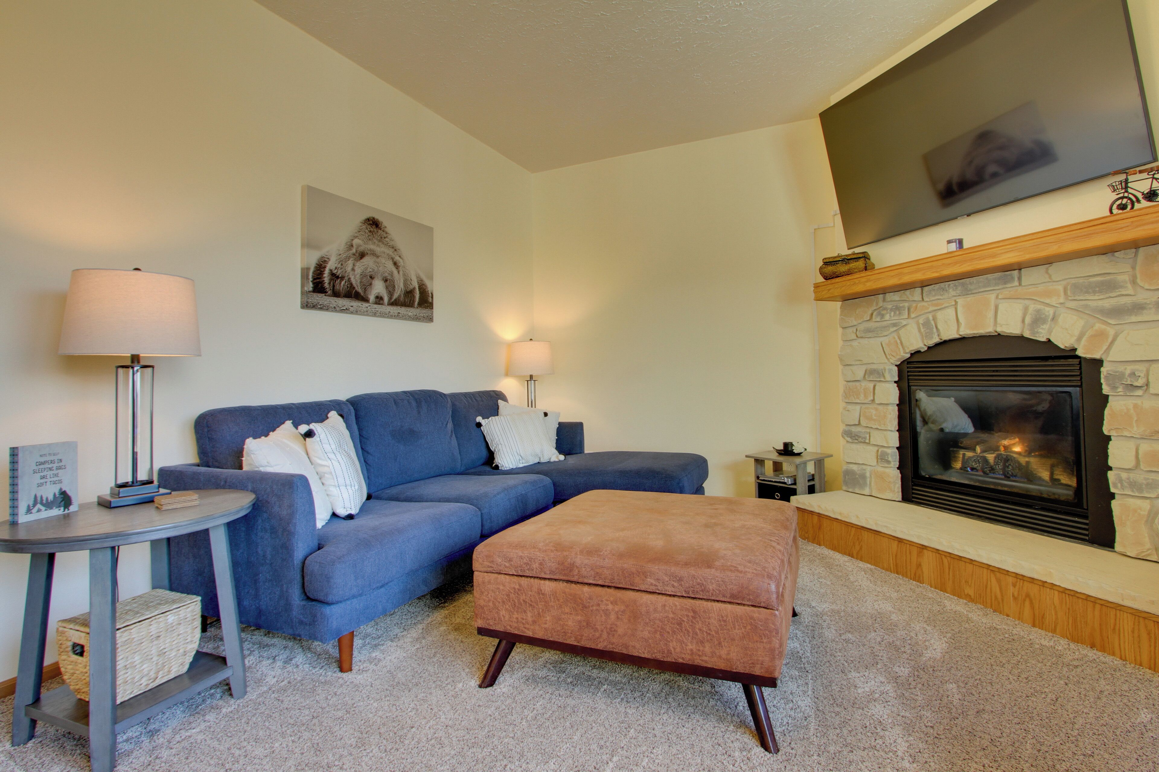 A living room with a blue sofa, a round side table, a small ottoman, a fireplace, and a mounted TV.