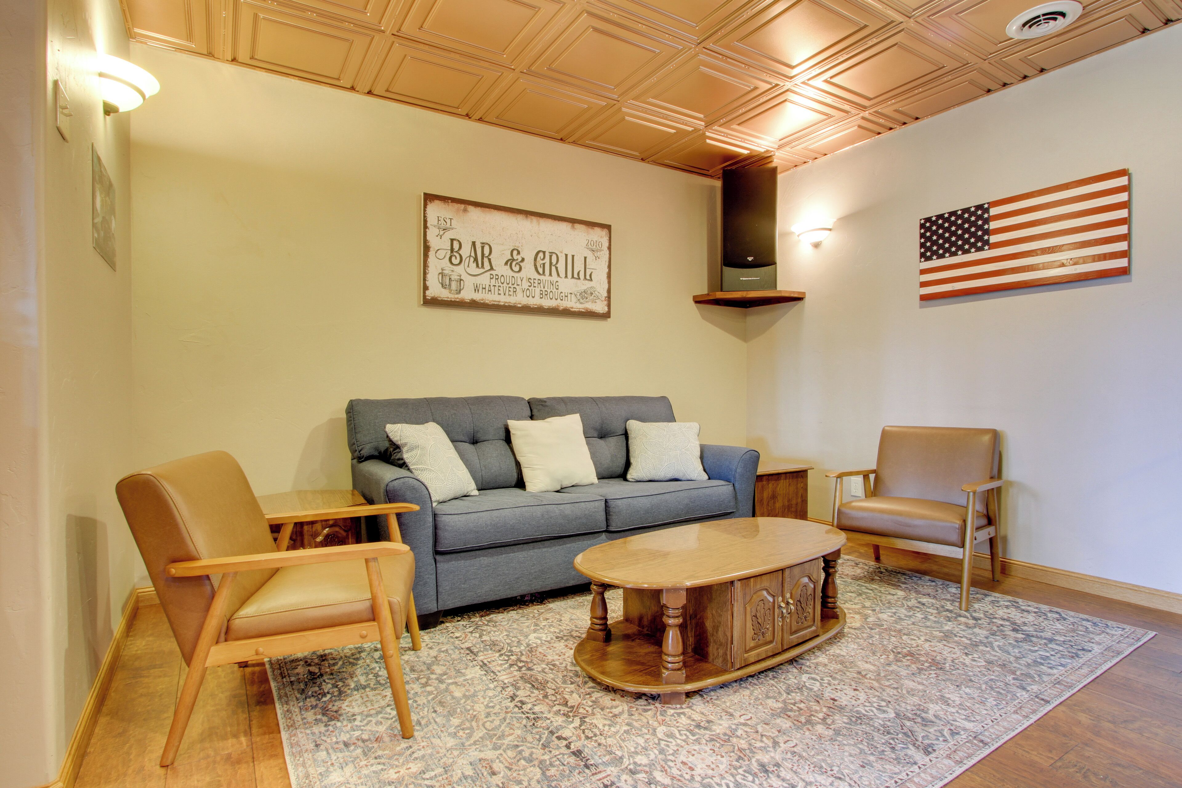 A living room with a sofa, two armchairs, a coffee table, and a rug. There is a sign on the wall that reads "BAR & GRILL" and an American flag.