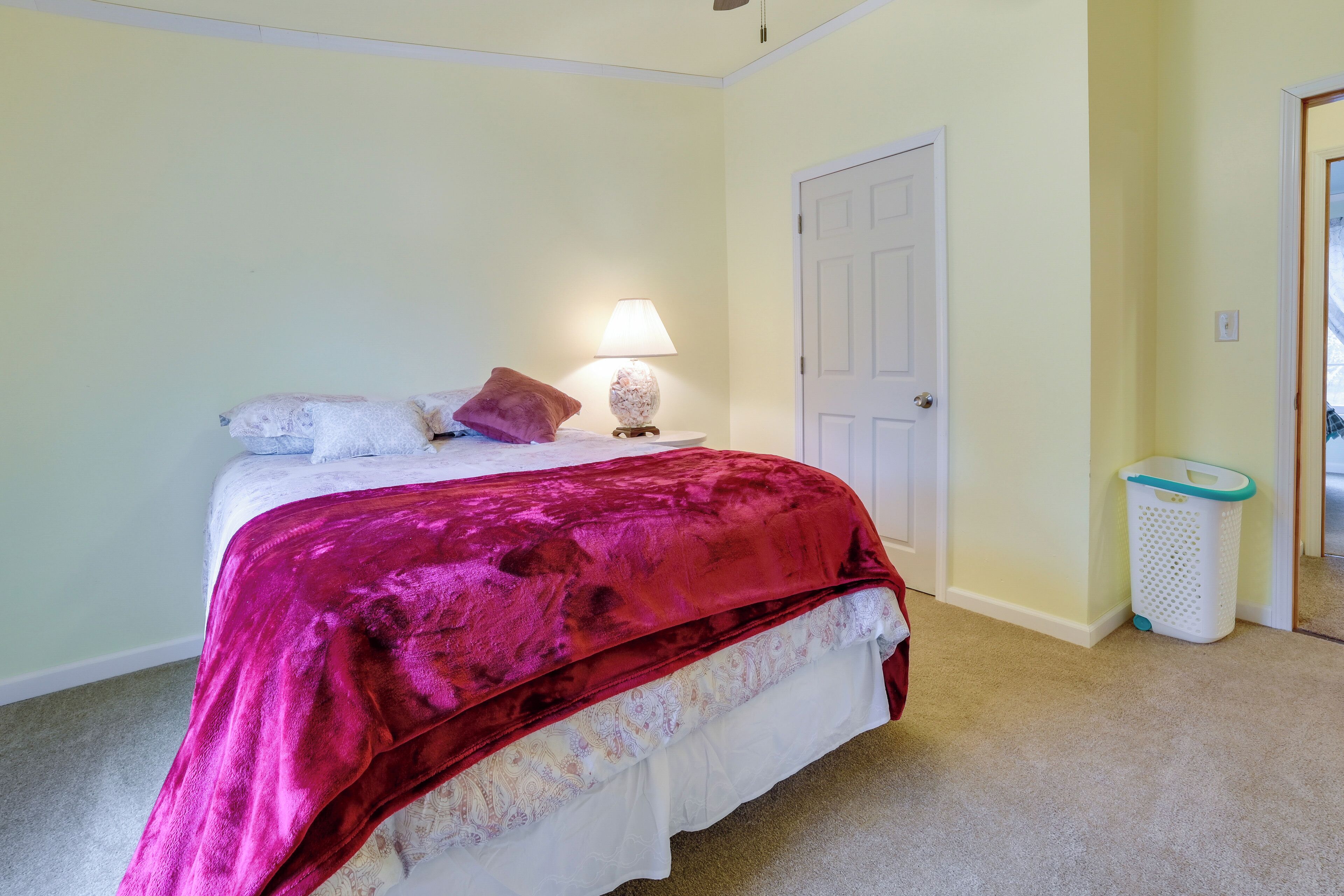 A bedroom with a bed, a nightstand, a lamp, a door, and a trash bin.