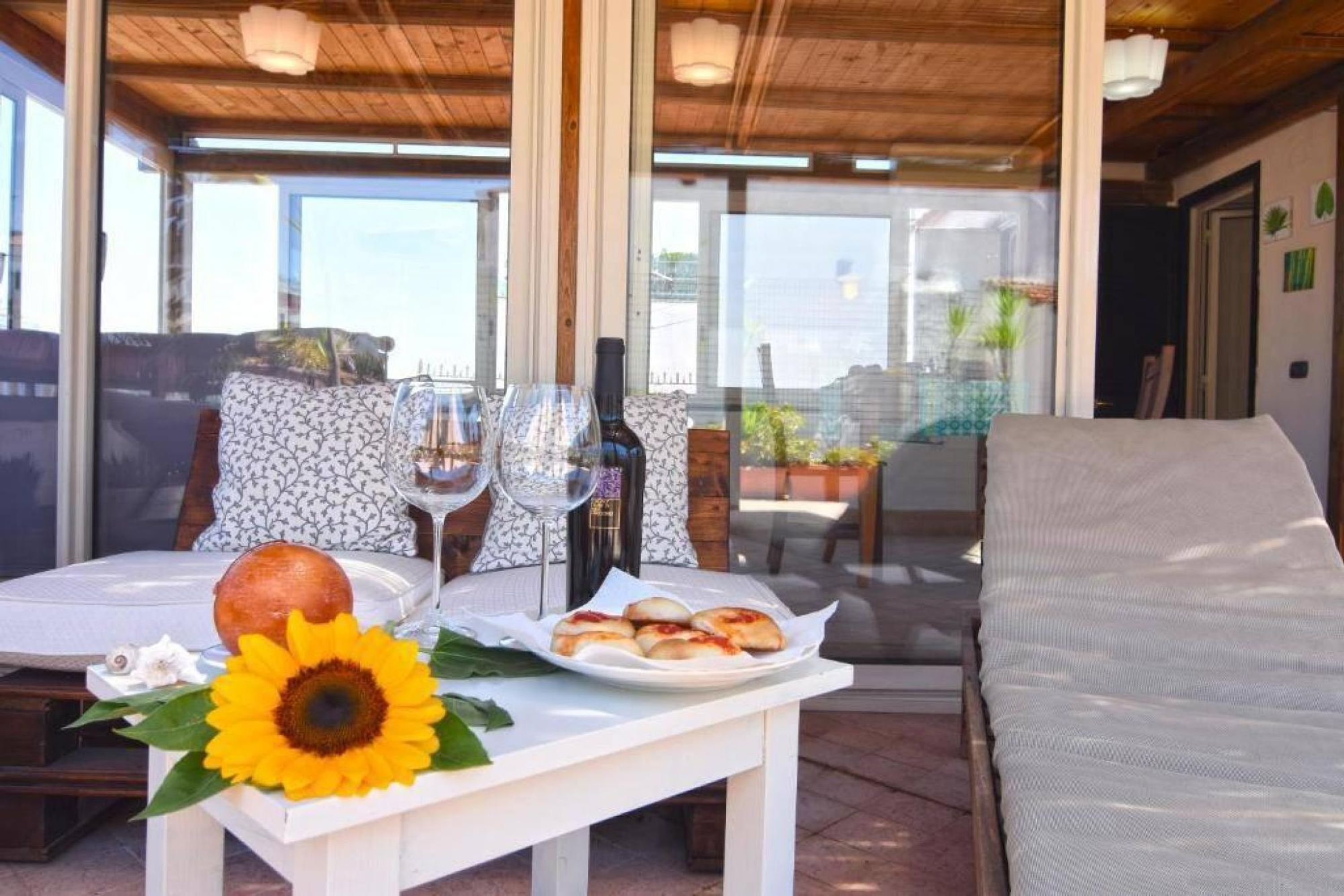 An outdoor dining area with a table set for two, featuring a plate of food, a bottle of wine, and a sunflower decoration.