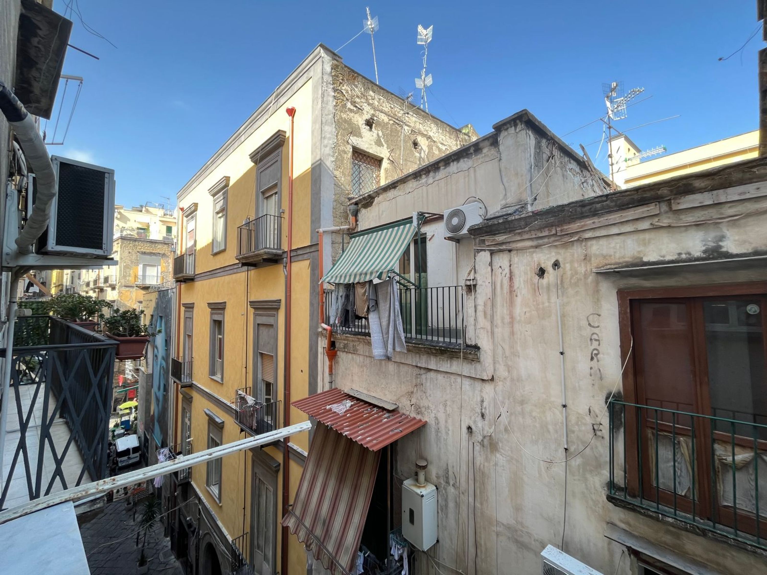A narrow urban street with closely packed buildings, some with balconies and air conditioning units.