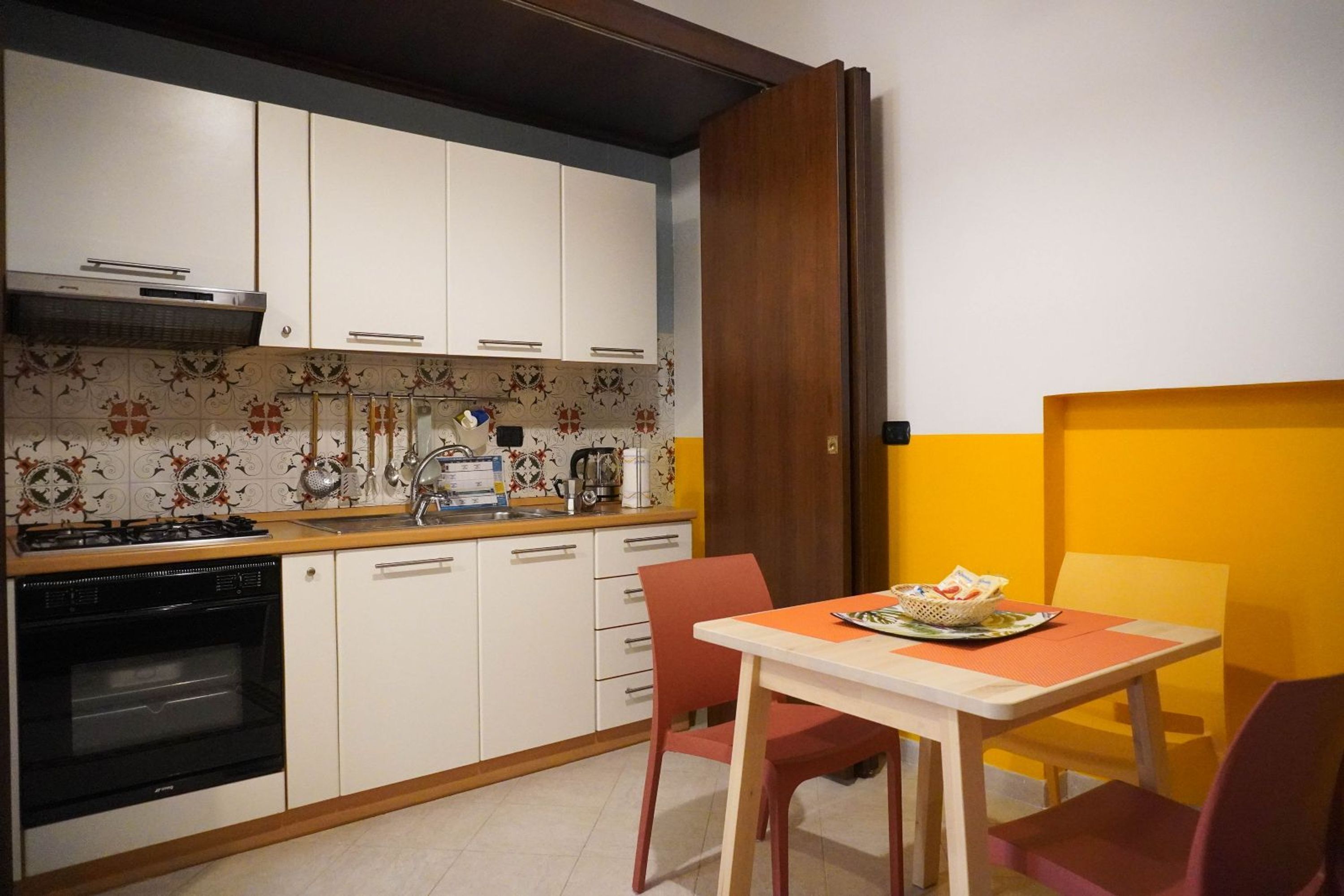 A compact kitchen with white cabinets, a stove, and a sink. There is a dining table with chairs and a basket of magazines.