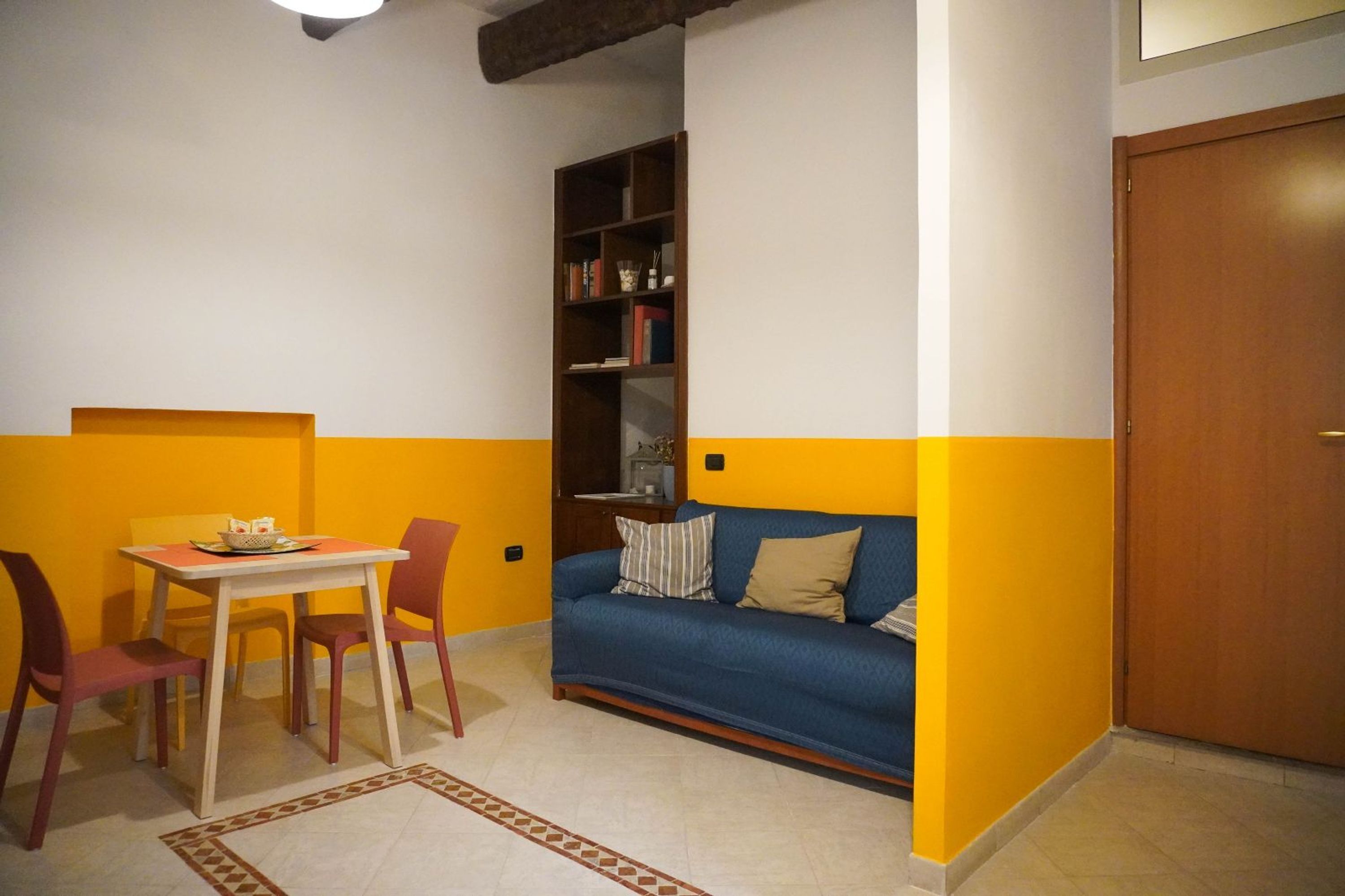 Un petit salon avec un canapé bleu, une bibliothèque en bois, une table à manger avec des chaises et un tapis à motifs.