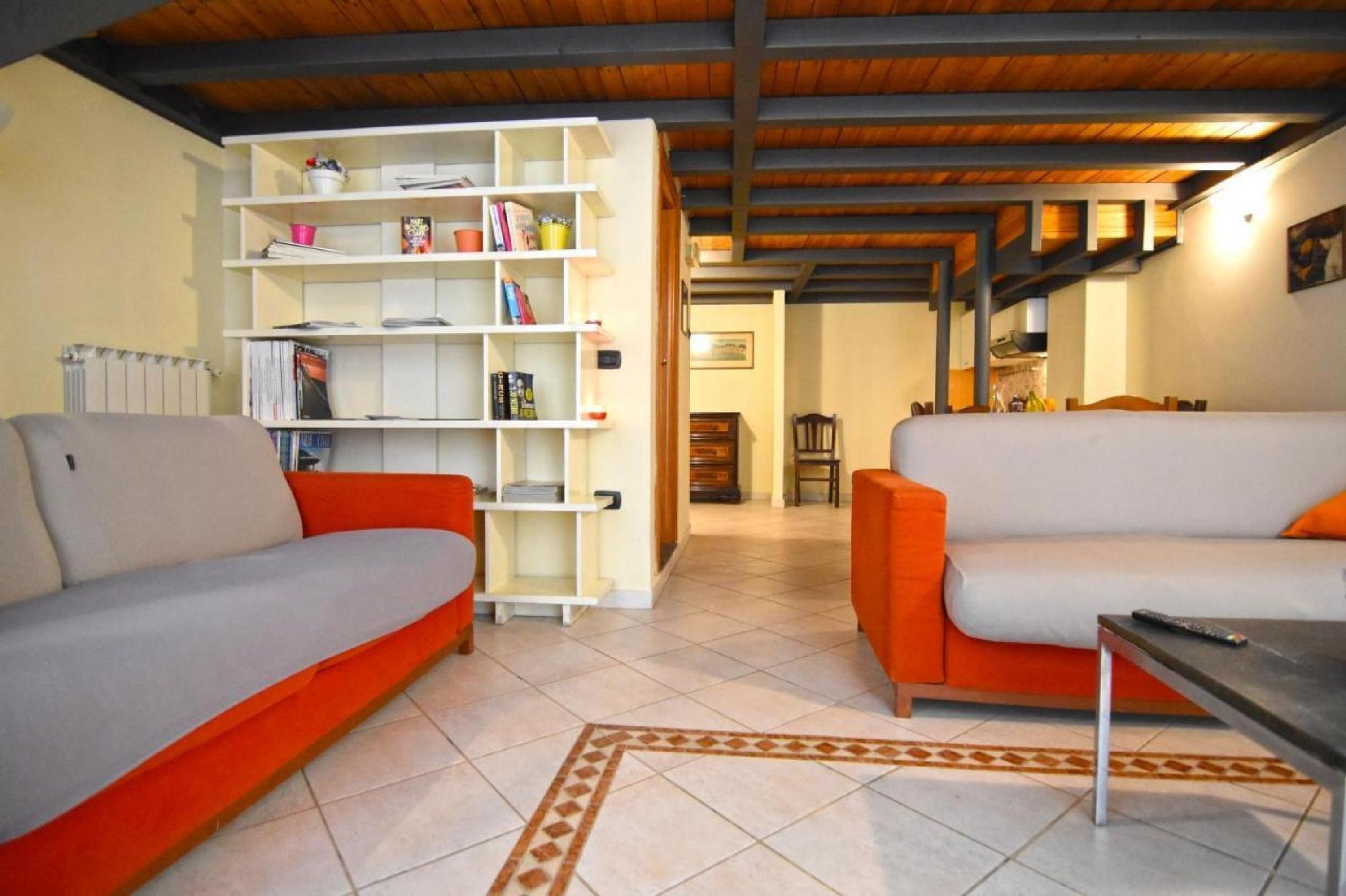 A living room with two orange sofas, a white bookshelf, and a wooden ceiling.