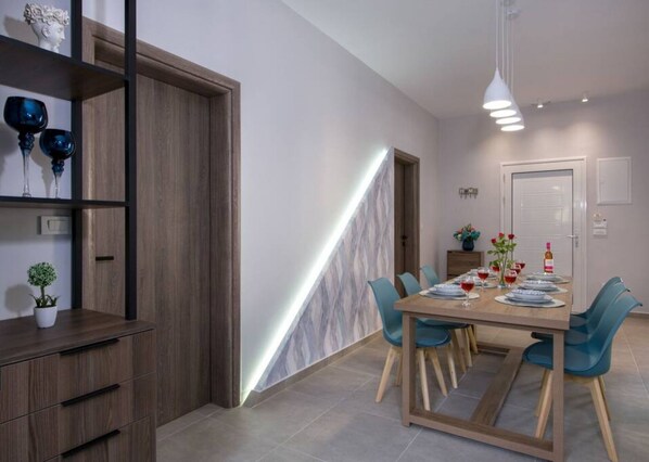 A modern dining area with a wooden table set for four, blue chairs, and a pendant light above. - Villa Eva Mari (Chania)