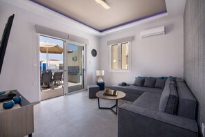 A modern living room with a grey sofa, a coffee table, and a dining area with chairs and an umbrella. - Villa Eva Mari (Chania)
