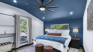 Un dormitorio con cama, lámparas de noche, ventilador de techo, ventana con cortinas y una mesita con lámpara.