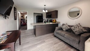 A modern living room with a grey sofa, a dining table with magazines, a kitchen area with dark cabinets, and a round mirror on the wall. - Lock Haven Grandview Hotel (Mill Hall)