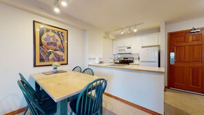 Un comedor con mesa de madera y sillas color turquesa, una cocina con armarios blancos y un refrigerador, y una puerta de madera.