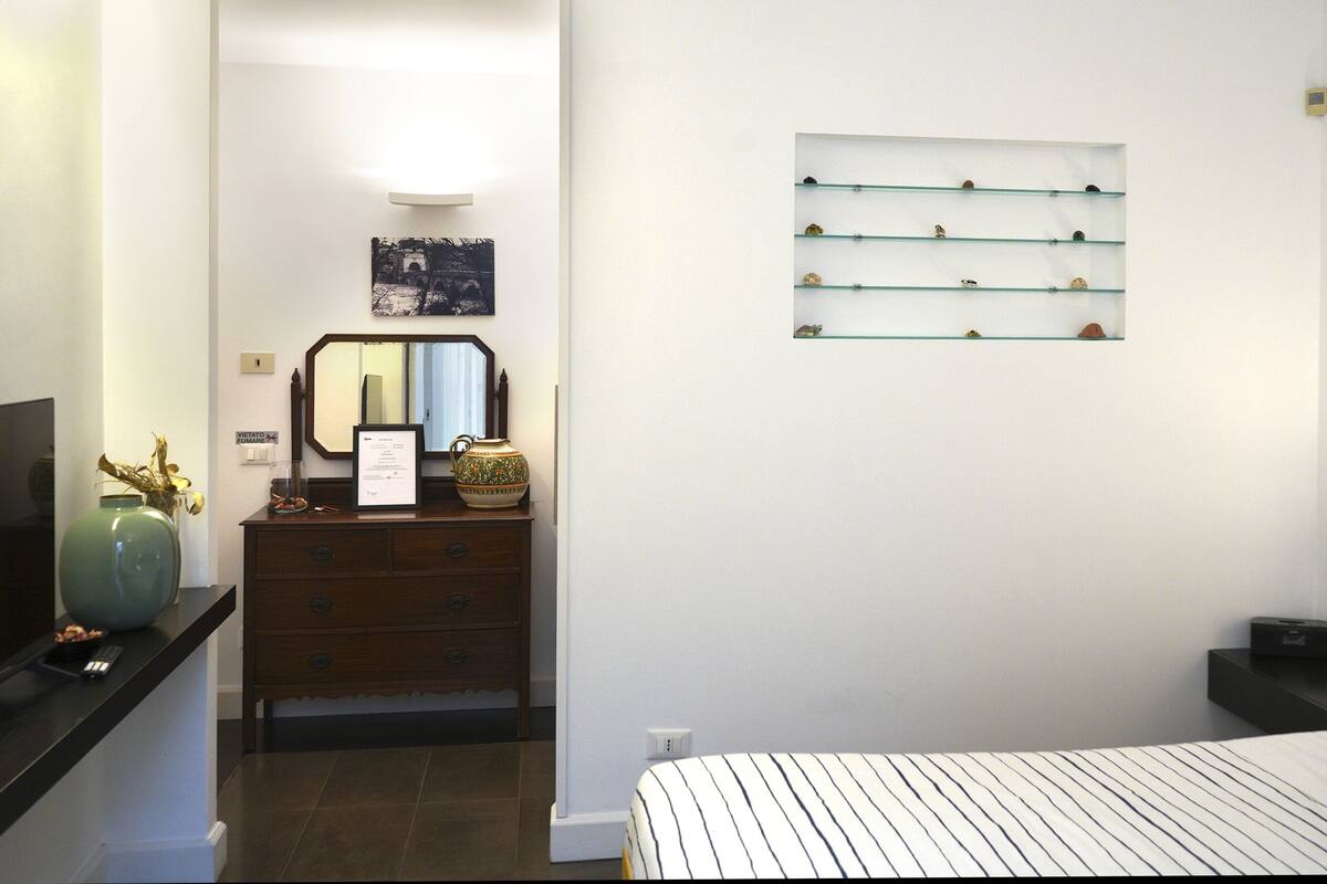 A bedroom with a bed, bedside table, lamp, and a wall-mounted shelf with decorative items.