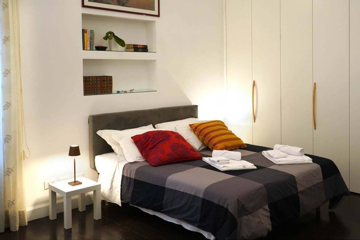 A neatly arranged bedroom with a bed, bedside tables, a wardrobe, and a shelf with books and a plant.
