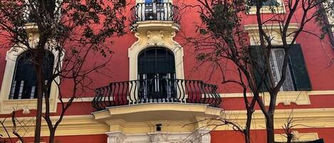 Um edifício com fachada vermelha, varanda com guarda-corpo preto e uma grande porta de madeira.