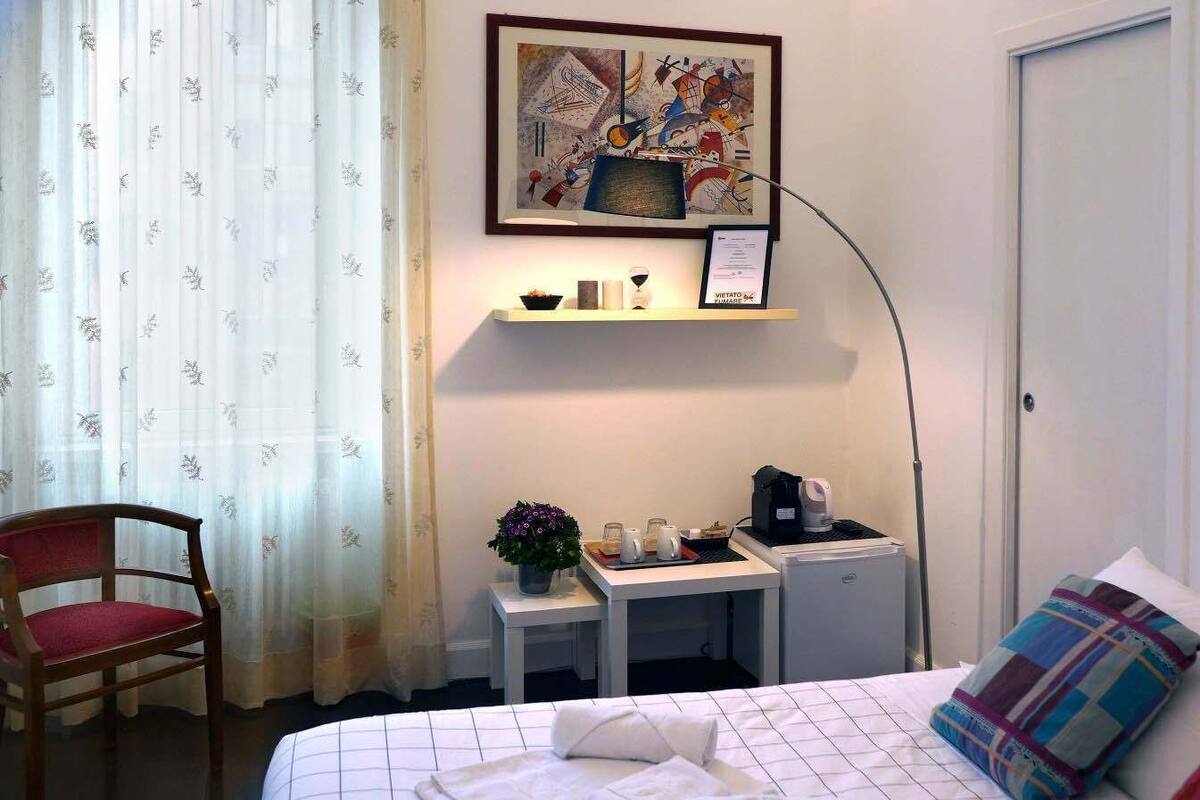 A hotel room with a bed, a desk, a chair, a coffee machine, and a framed artwork on the wall.