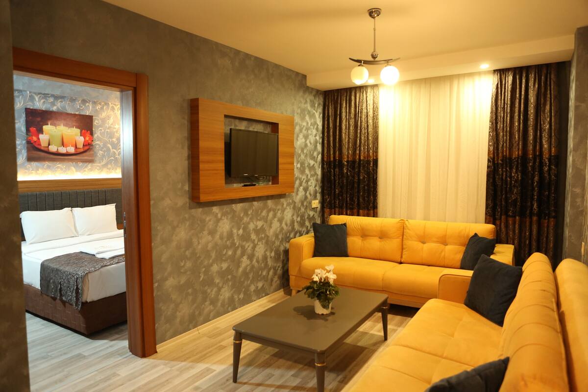 A modern living room with a sofa, a coffee table, a TV mounted on the wall, and a bed in the adjacent room.