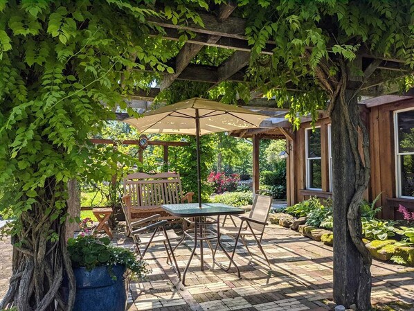 An outdoor patio with a table and chairs, a pergola with an umbrella, and lush greenery.