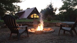 Uma área de estar rústica ao ar livre com cadeiras Adirondack ao redor de uma lareira, com uma cabana de madeira ao fundo.