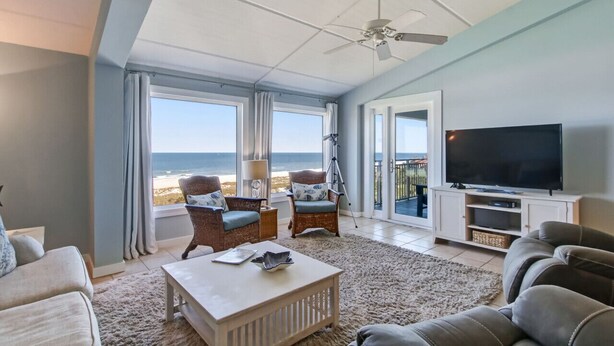 A spacious living room with a large TV, comfortable seating, and a balcony with a view of the beach.