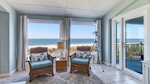 A room with a view of the beach, wicker chairs with cushions, a lamp, and a camera tripod.
