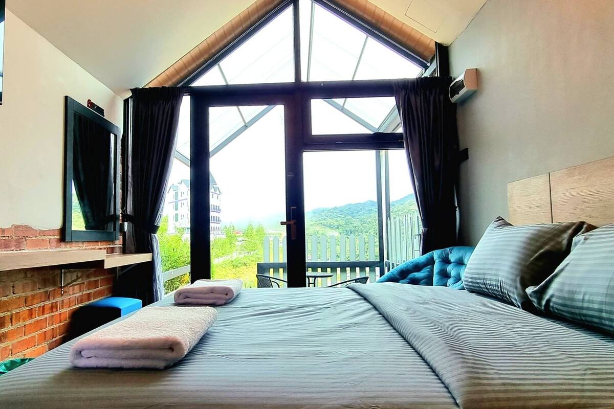 A hotel room with a large bed, a TV on the wall, and a view of greenery outside.
