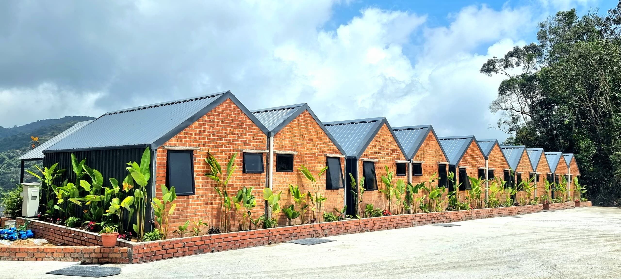 A row of brick buildings with triangular roofs, surrounded by greenery and a clear sky.