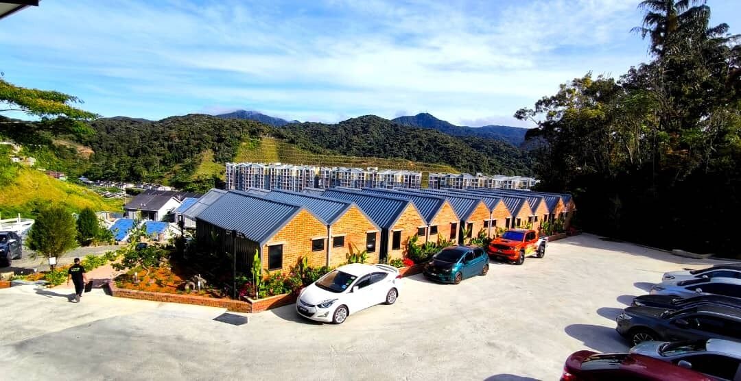 Un hotel con una fila di stanze dai tetti blu, un parcheggio con diverse auto e montagne sullo sfondo.