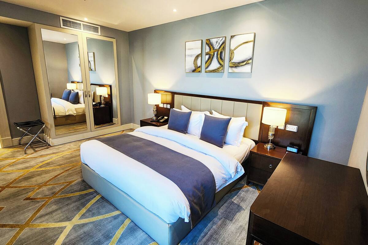 A hotel room with a large bed, a nightstand with a lamp, a mirror, and artwork on the wall.