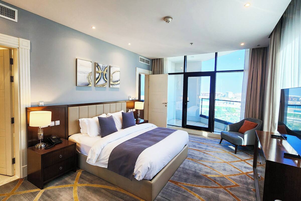 A hotel room with a large bed, a desk with a lamp, a chair, and a large window with a city view.