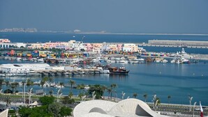 A coastal cityscape with colorful buildings, a marina with boats, and a distinctive white curved structure in the foreground. - Sherao Hotel Doha - Next to National Museum of Qatar & Metro (Doha)