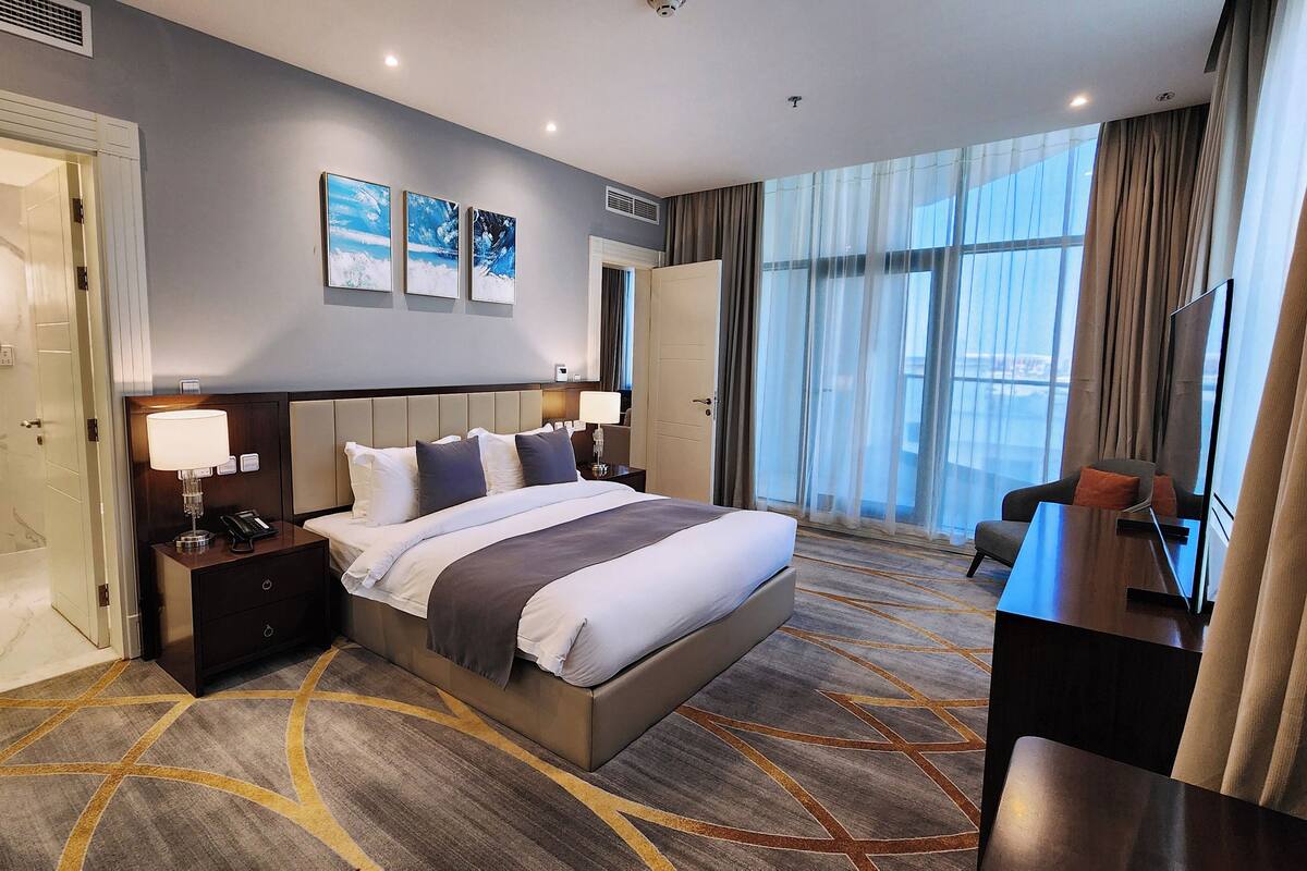 A hotel room with a large bed, bedside tables, a desk, a chair, a bathroom with a mirror, and artwork on the wall.