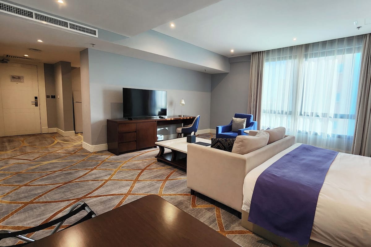 A hotel room with a bed, a sofa, a desk with a TV, and a large window with curtains.