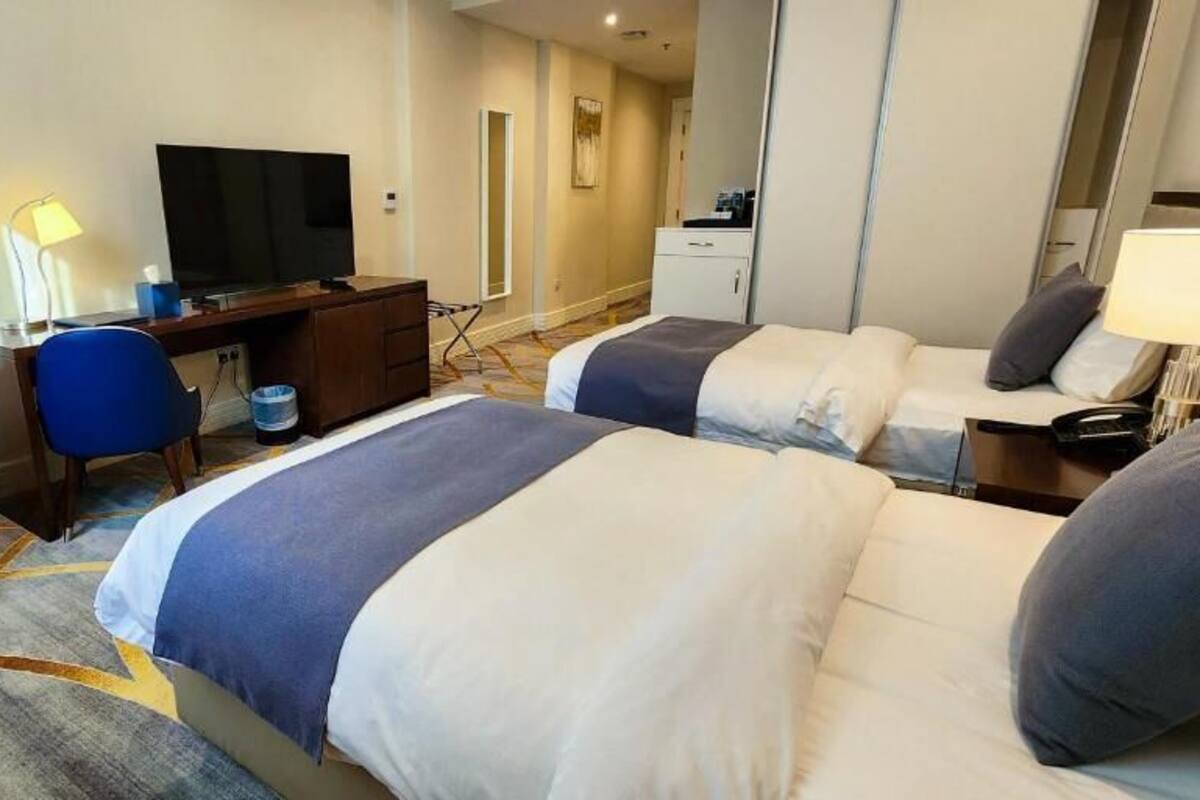 A hotel room with two beds, a desk with a TV, a chair, and a small table with a lamp.