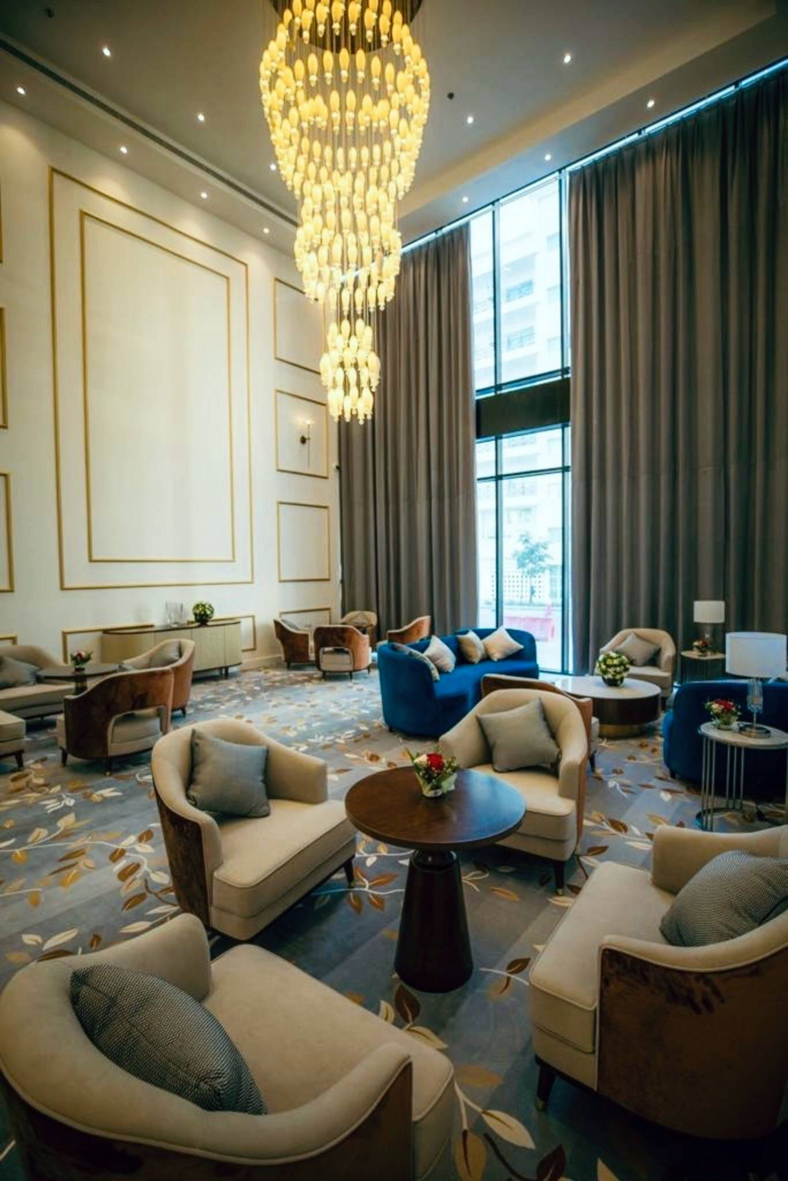 Un hall d’hôtel spacieux, agrémenté d’un grand lustre, de plusieurs espaces aménagés pour s’asseoir et de grandes fenêtres avec des rideaux.