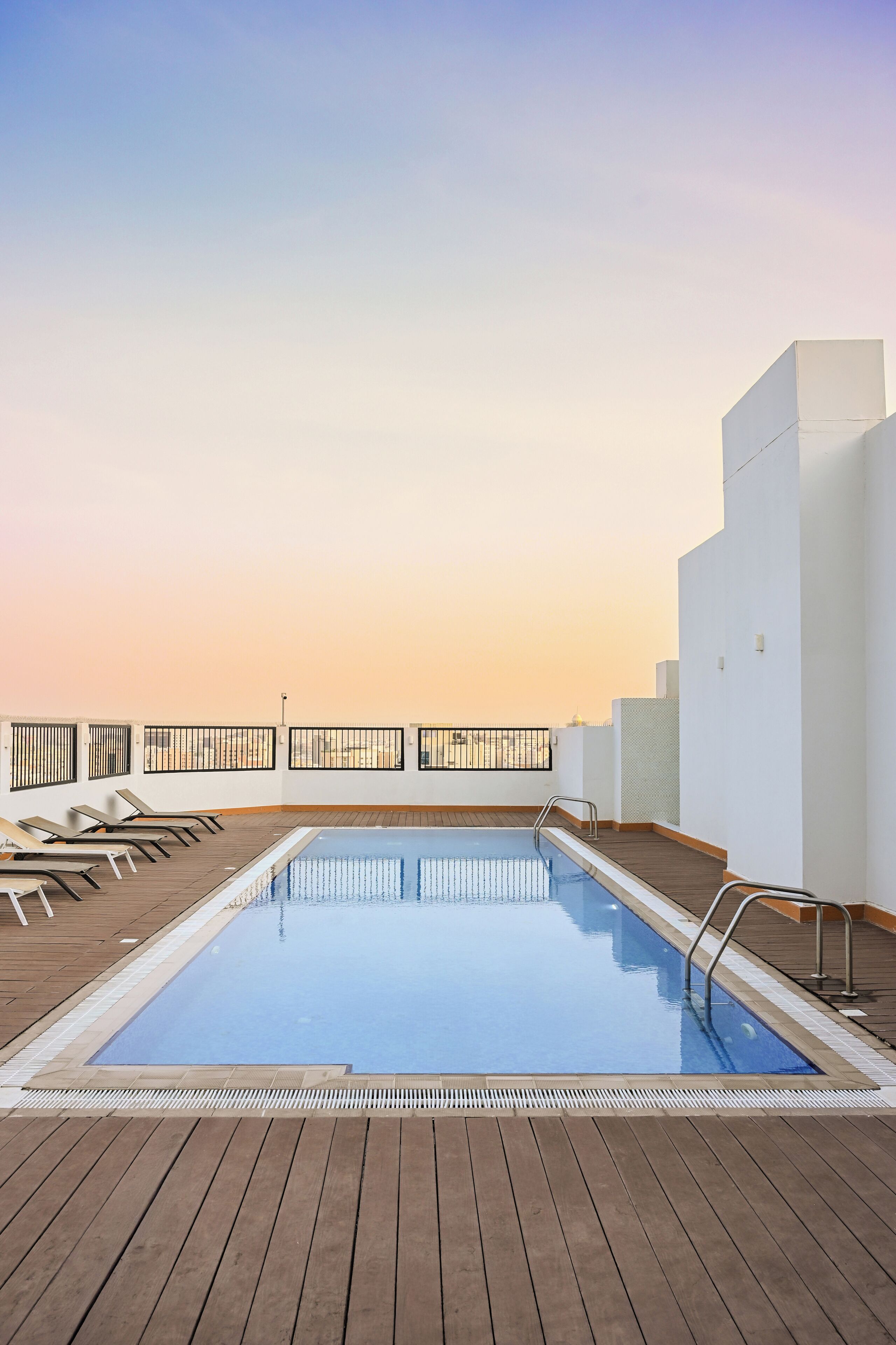 A rooftop pool with a wooden deck and lounge chairs, surrounded by a white wall and a metal railing.