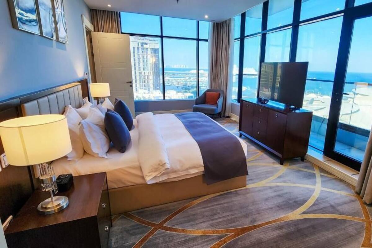 A hotel room with a large bed, a TV, a desk with a lamp, and a view of the city through large windows.