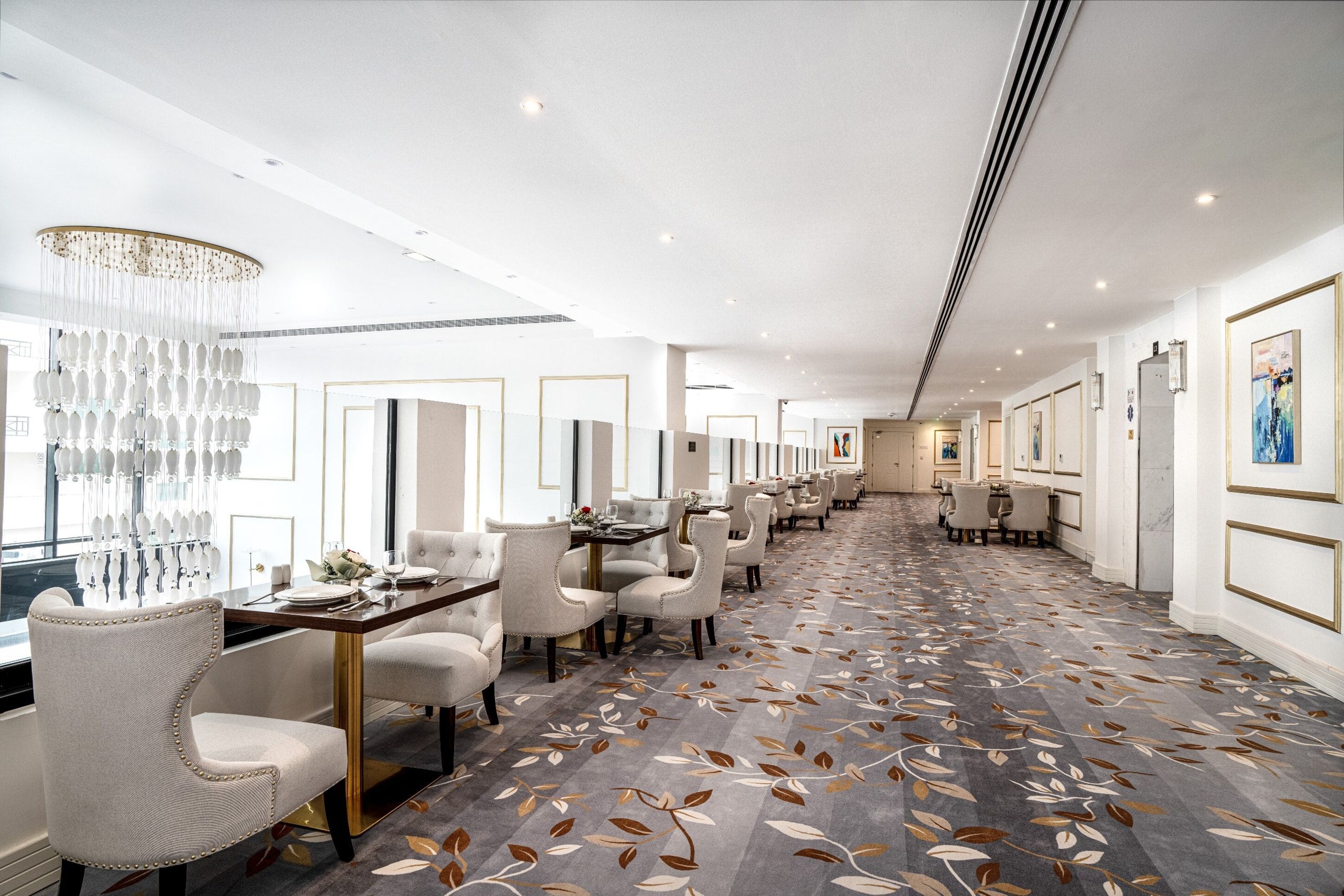 A modern hotel lobby with a long reception desk, white upholstered chairs, a patterned carpet, and a chandelier.
