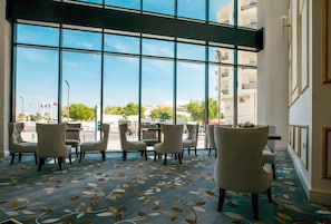 Un hall de hotel con ventanas amplias, alfombra estampada y varias sillas dispuestas para sentarse.