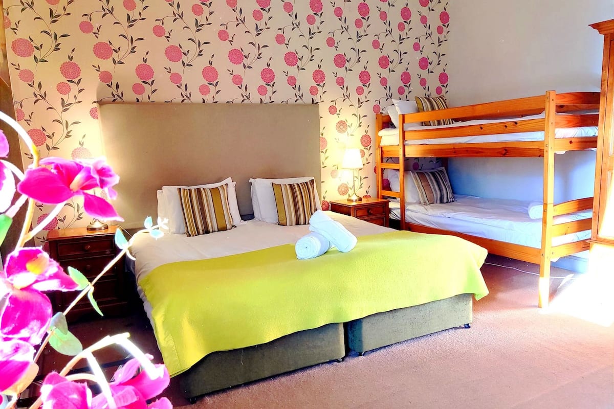 A bedroom with a bunk bed, a large bed with pillows, a bedside table with a lamp, and floral wallpaper.