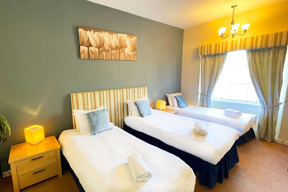 A hotel room with three single beds, a nightstand with a lamp, a wall-mounted artwork, and a window with curtains.