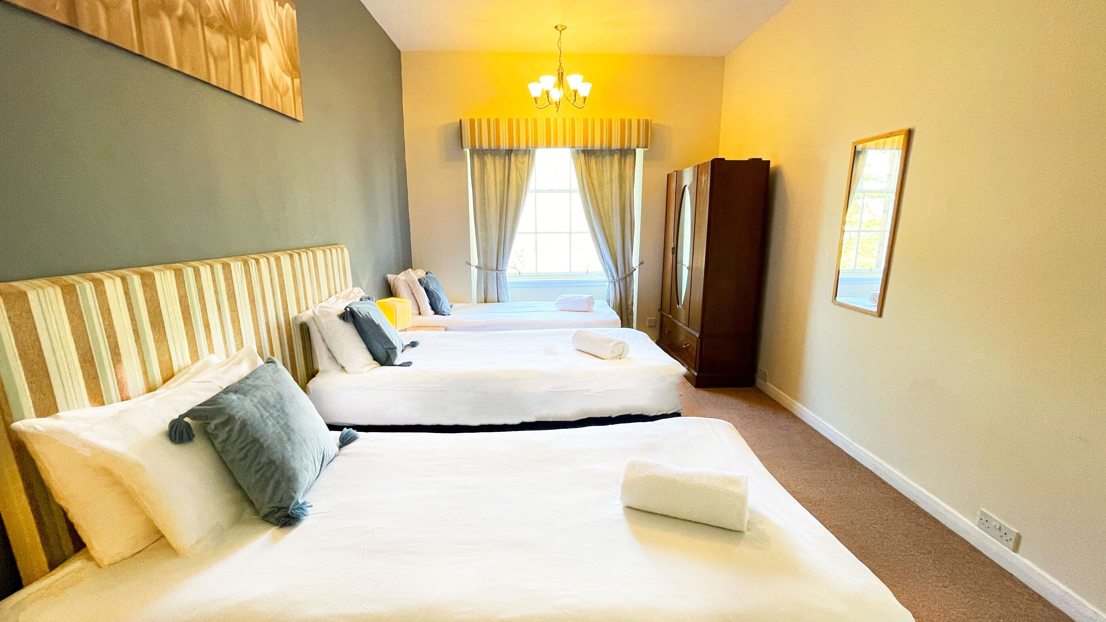 A hotel room with two beds, a wardrobe, a chandelier, and a window with curtains.