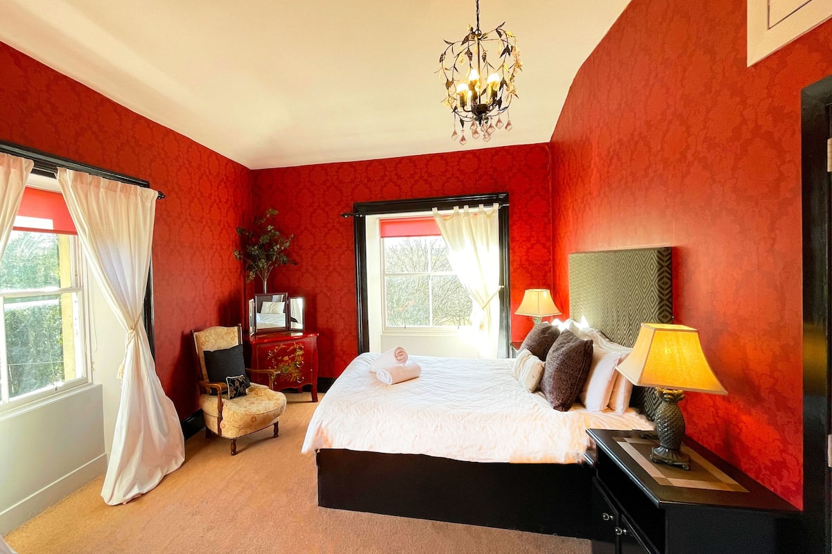 A bedroom with a bed, a nightstand, a chair, a window with curtains, and a chandelier.