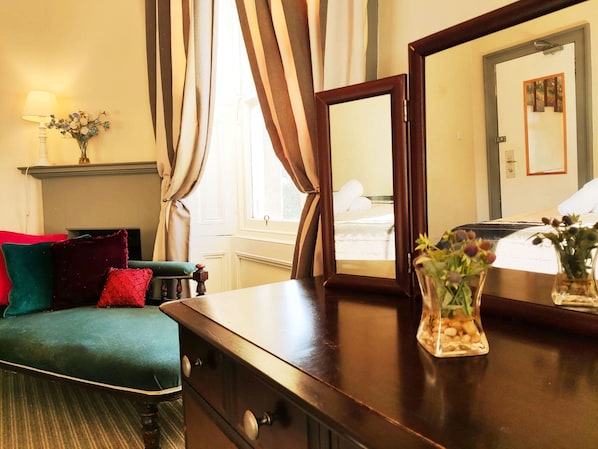 A hotel room with a wooden dresser, a mirror, a bed with white linens, a green sofa with red and teal cushions, a vase with flowers, and a window with curtains. - Kirkhill Estate Rooms (Gorebridge)